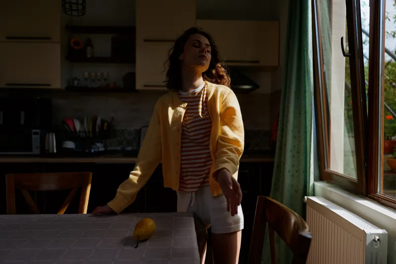 Young woman stands in a cozy home kitchen beside a table with a lemon, wearing a yellow cardigan and striped top, bathed in natural light creating a casual lifestyle scene