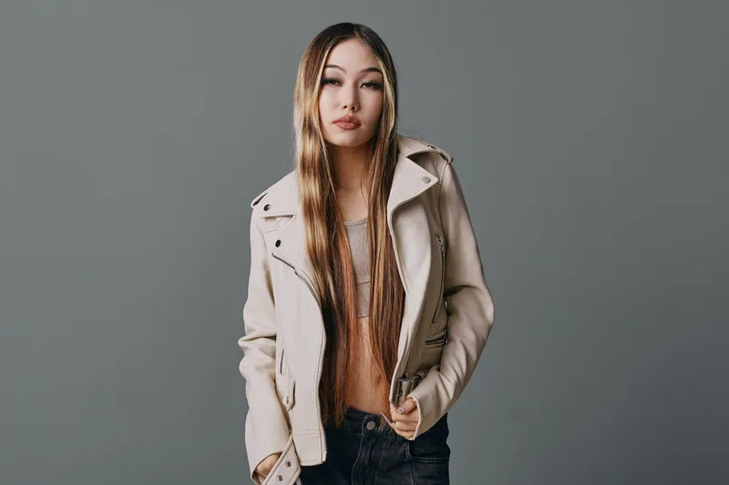 Young woman with long straight hair wearing beige jacket and casual clothes standing against gray background with relaxed confident expression and hands in pockets.