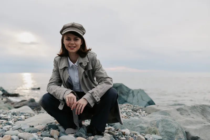 Young woman in casual outfit and hat sits on pebble beach near calm sea under cloudy sky. Natural light highlights relaxed mood and outdoor lifestyle concept.