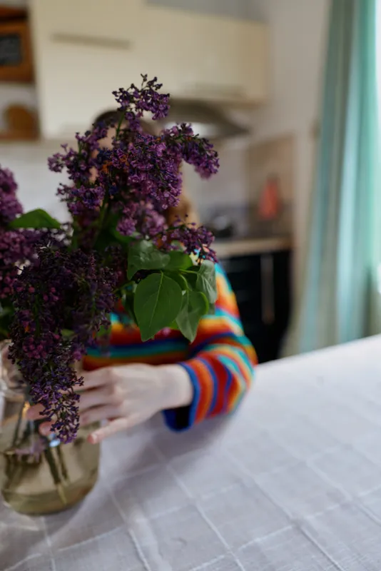 Woman indoors, holding a bouquet of lilac flowers, striped sweater visible, soft natural light, cozy kitchen scene, relaxed pose, fresh floral centerpiece, warm home mood, bright colors and tender