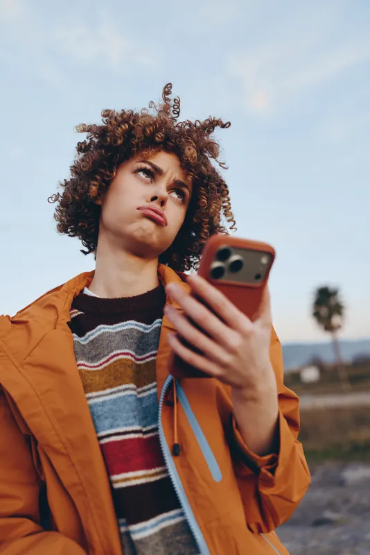 Woman lifestyle portrait outdoors, curly hair and orange jacket, holding smartphone with screen visible, looking at camera, soft sunlight and blue sky, calm travel mood