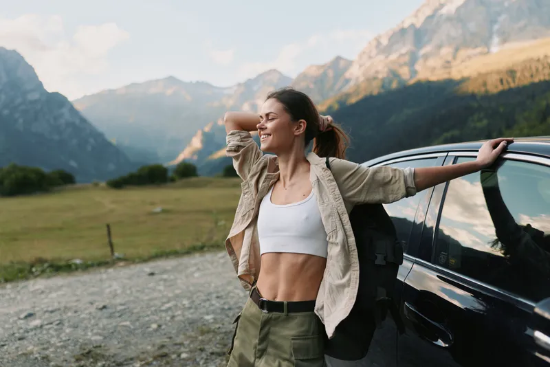 Happy young woman enjoys nature standing near car with mountains in background, dressed in casual outfit, lifestyle concept of outdoor adventure and freedom.