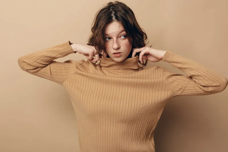 Young woman with short brown hair wearing beige knitted sweater pulls collar up. Neutral background and soft lighting create cozy autumn fashion concept with casual style.