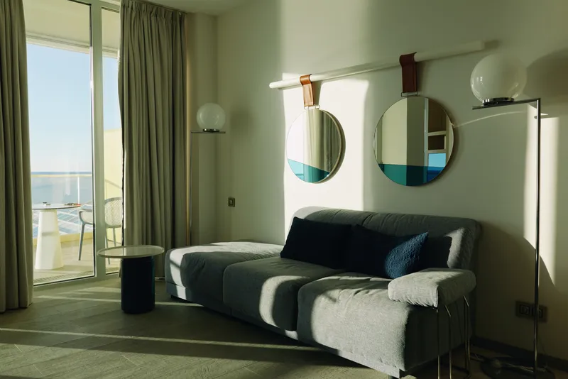 Living room with a modern sofa and round mirrors, bathed in natural light from a balcony window. Cozy seating, sleek design, soft textures and stylish home interior.