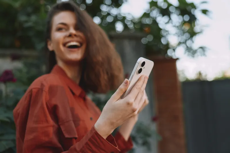 Cheerful woman outdoors, laughing with phone in hand, casual jacket, sunlit moment, relaxed posture, natural colors, lush garden background