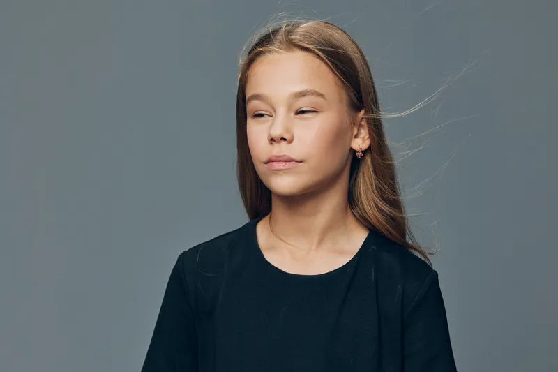 Girl with long hair wearing black shirt looking sideways with calm expression on gray background in studio lighting. Youth, child and casual portrait with natural hairstyle.