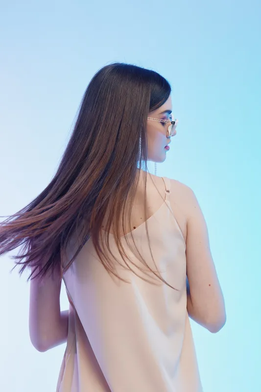 Young woman with long hair wearing glasses and a beige sleeveless top looks sideways in a studio with blue gradient background. Concept of beauty, fashion, and style.