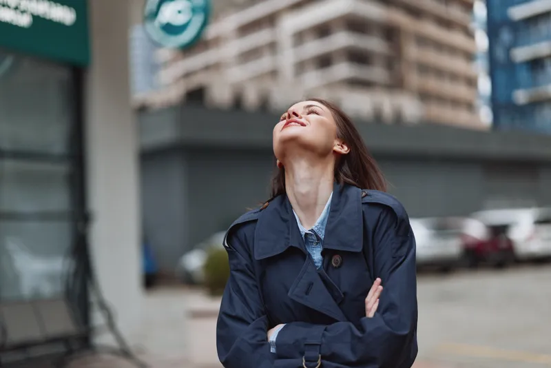 Young woman with long brunette hair wearing a navy trench coat stands outdoors in an urban setting with her eyes closed and head tilted back, enjoying the moment.