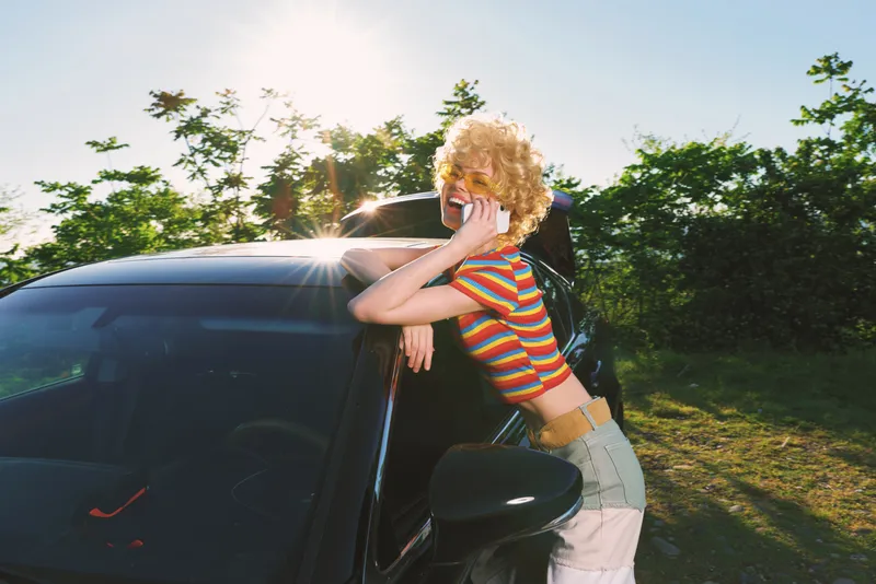 Woman leans on a car, smiling, striped shirt, sunny outdoor setting, retro film color aesthetic, casual lifestyle shot for editorial use. Bright light and warm tones enhance mood. Casual vibe.