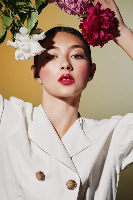 Young woman with vivid makeup and glossy red lips poses with fresh flowers near her head wearing white fashion outfit. Clean style with soft shadows on beige background.
