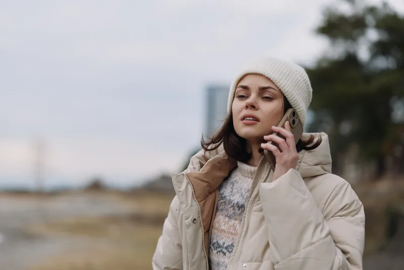Woman on a mobile phone outdoors in a warm beige jacket and knit beanie, mid conversation in a calm winter setting, soft light, casual style and relaxed mood, subtle urban vibe with distant blurred