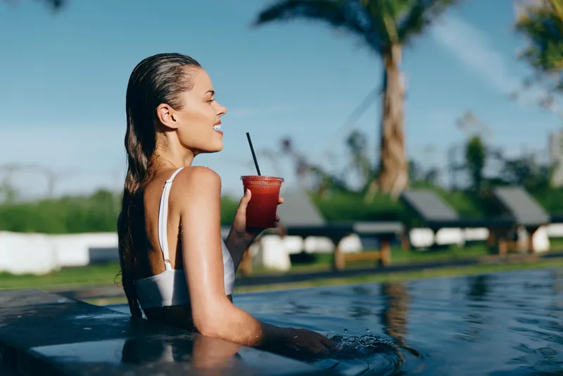 Poolside scene featuring a woman in a white swimsuit sipping a red tropical drink, sunlit water glistening, relaxed posture, and palm trees in the background for a carefree summer mood