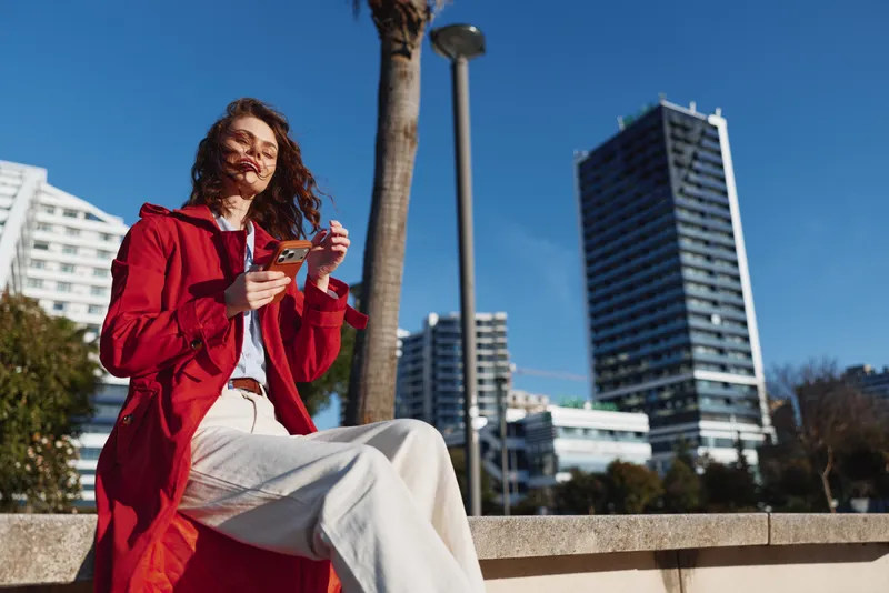 Young woman with curly hair wearing a red coat sits on a stone bench using her smartphone on a sunny day in an urban environment with tall buildings and palm trees.