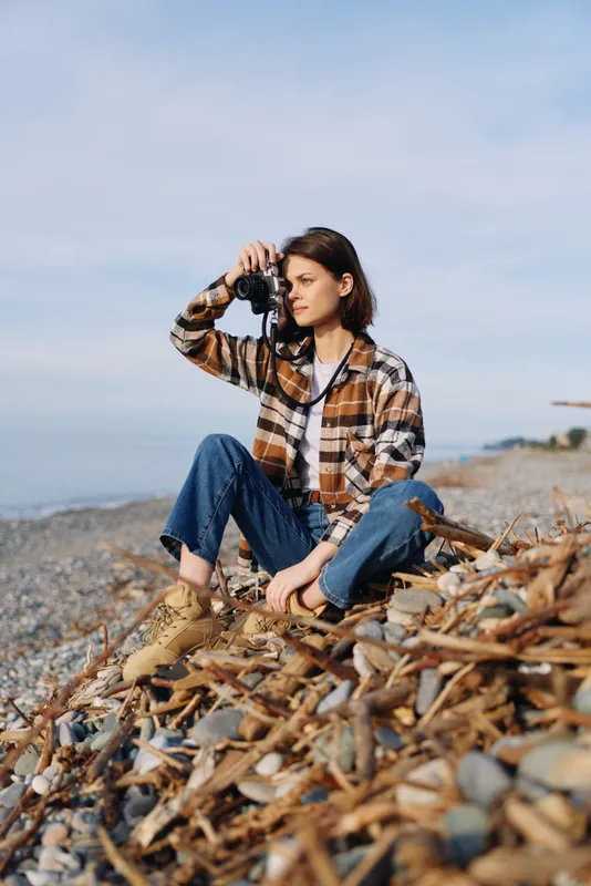 Photographer, camera, beach, outdoors, casual, seascape, landscape, nature, posing, shoreline enjoying a quiet moment as rocks and driftwood frame the scene during daylight.