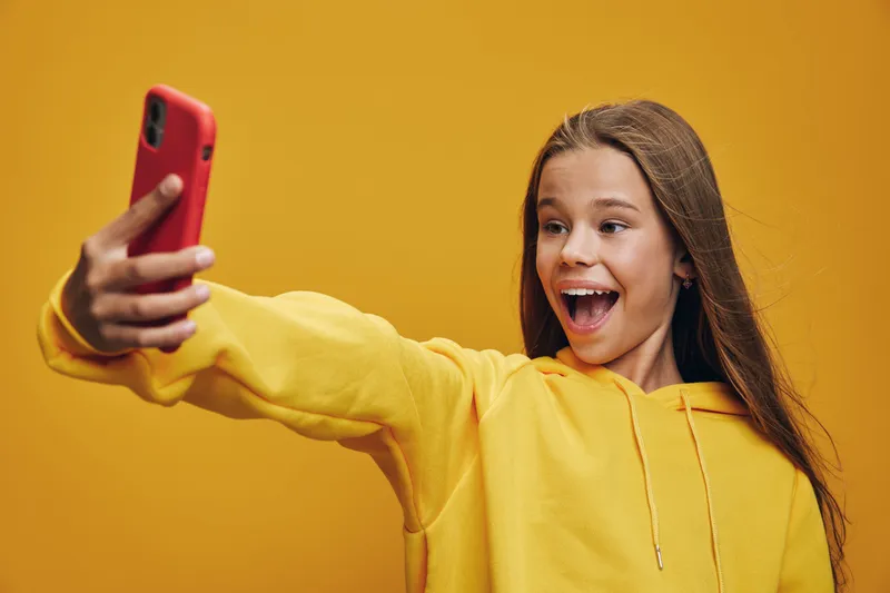 Girl in yellow hoodie takes a selfie on smartphone with excited expression against yellow background. Child with long hair and happy face capturing photo indoors with mobile phone.