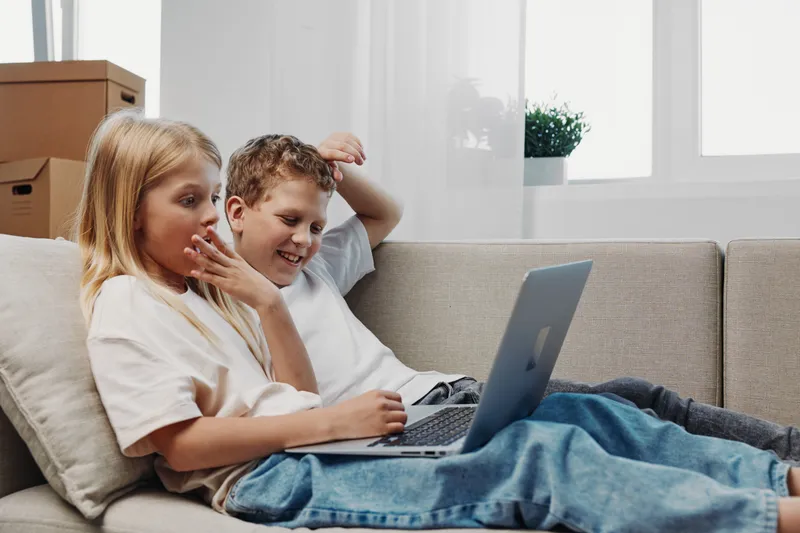Two children relax on a couch using a laptop, focused and engaged in a cozy home environment with natural light.