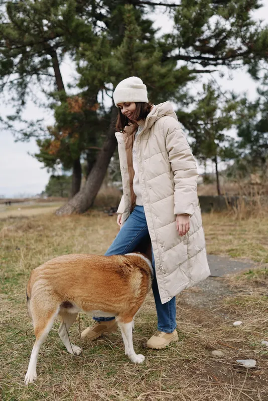 woman and dog outdoors in a warm coat walking along a grassy path with trees in the background, casual lifestyle moment capturing fresh air and calm companionship