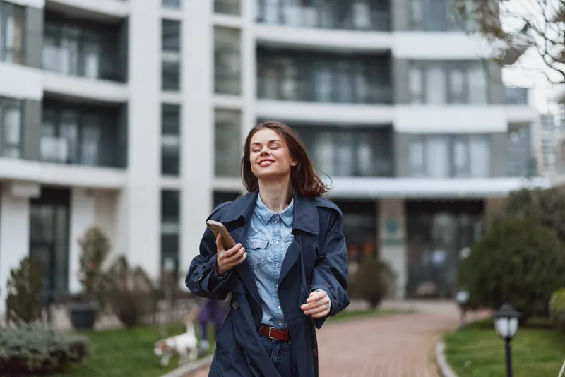 Young woman with denim jacket walks outside a modern building, holding a phone and smiling, conveying casual urban confidence.