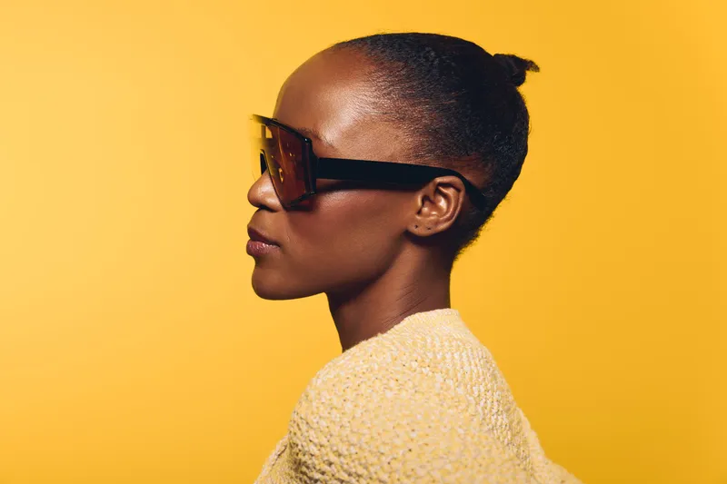 Profile of a confident dark skinned African woman wearing large sunglasses and a knitted sweater on yellow background. Concept of fashion, style, and modern beauty portrait.