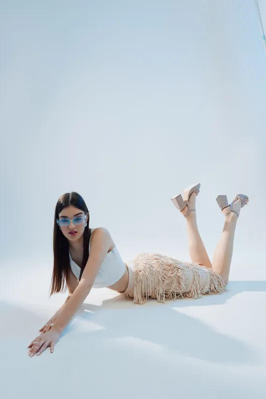 Young woman in a white top and fringe skirt poses on the floor wearing sunglasses and high heels. Studio shot with soft lighting. Concept of woman beauty fashion and style.