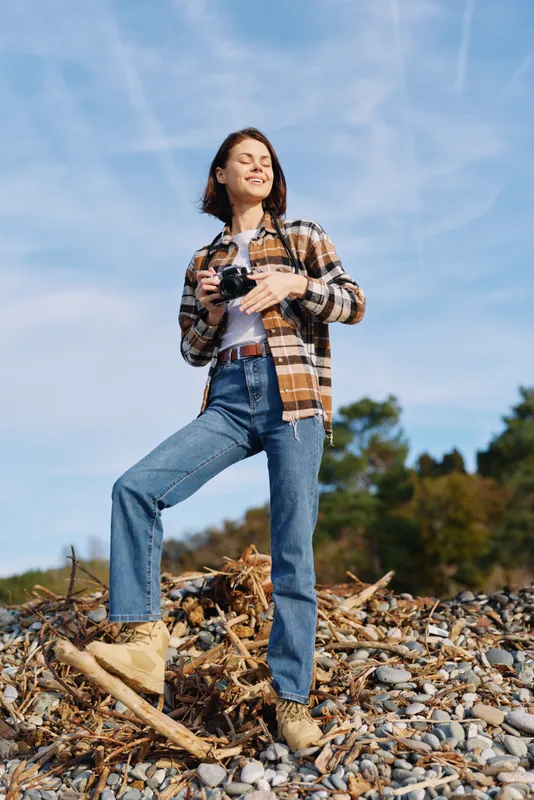 Woman stands confidently on rocky terrain under a bright blue sky, holding a camera and dressed in jeans and plaid shirt for an outdoors lifestyle photo