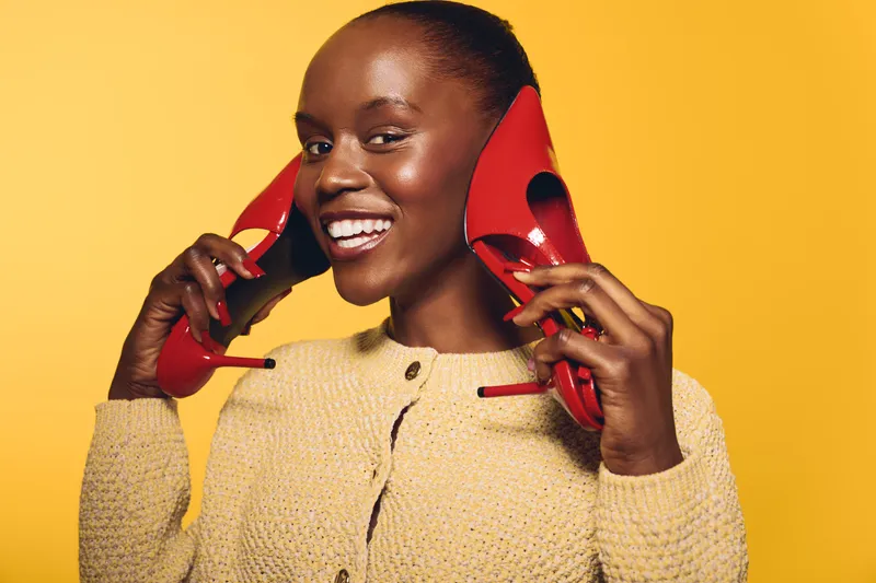 Smiling black African woman holds red high heels near face on yellow background. Happy dark skin female model wears beige sweater with positive expression. Concept for fashion and joy.