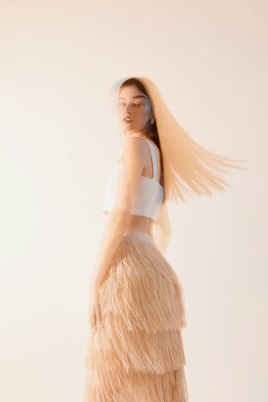 Young woman in a white crop top and fringed skirt poses with flowing hair in soft light. Concept for woman beauty fashion, elegance, and style in studio setting.