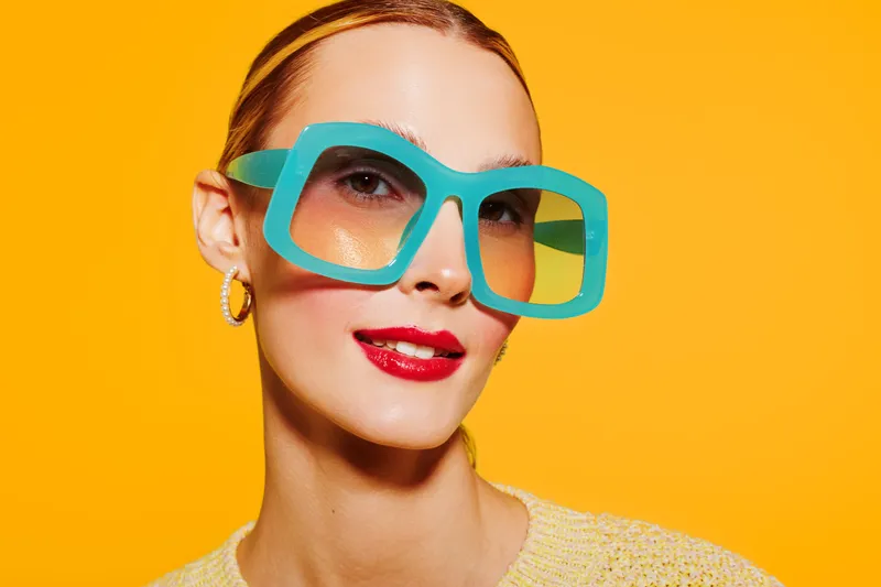 Close-up of a stylish woman wearing oversized turquoise sunglasses and red lipstick against a bright yellow background. Concept of fashion, summer, and vibrant style.