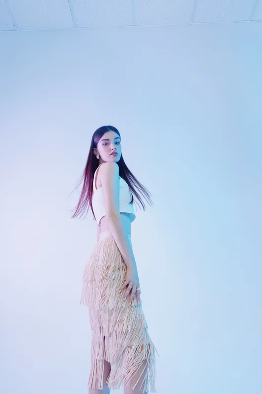 Young woman with long hair wearing a fringe skirt and white top poses confidently in a minimalist studio with soft blue lighting. Concept of beauty fashion and style.