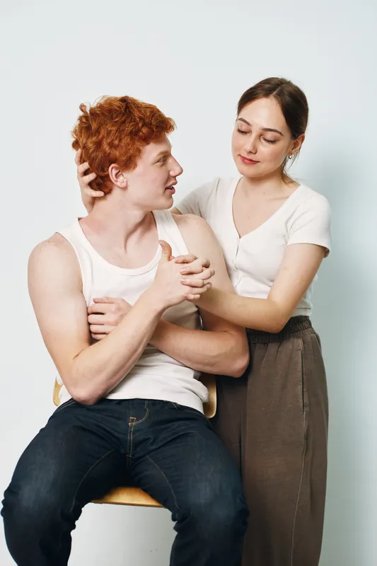 Young man and woman in casual clothes showing affection. Woman gently touching redhead man with arm around shoulder in relaxed indoor setting.