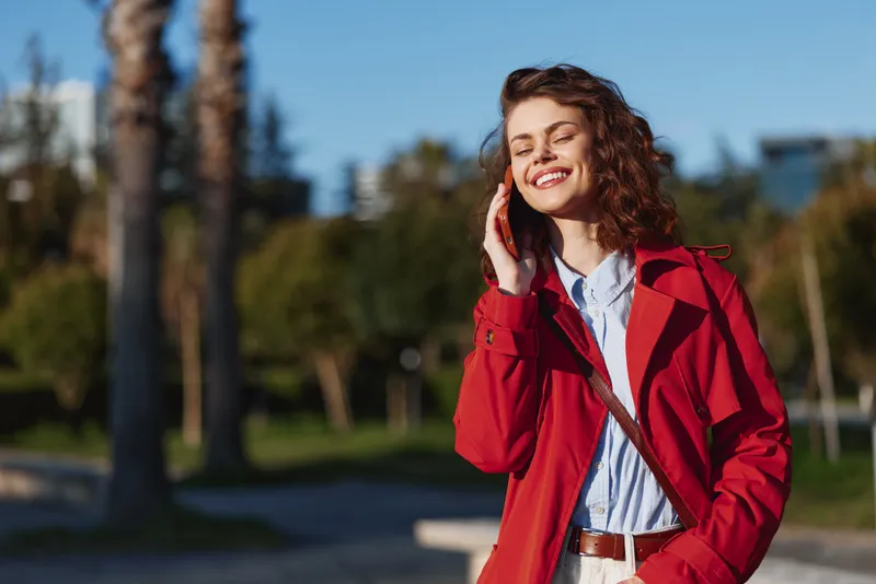 A young woman with curly hair wearing a red coat smiles while talking on a phone in a sunny park, conveying casual connection, confidence, and a candid outdoor moment.