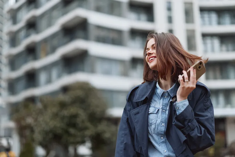 Young adult woman with brunette hair wearing a denim shirt and casual jacket talks on her smartphone in an urban street scene, smiling as modern architecture frames the shot.