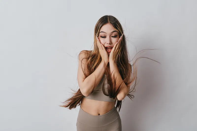 Young woman with long hair wearing beige activewear making excited surprised expression with hands on cheeks against plain light background. Fitness, emotion and casual lifestyle.