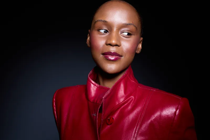 Portrait of a confident dark skinned African woman wearing a red leather jacket against a black background. Studio lighting highlights her smooth skin and thoughtful expression.