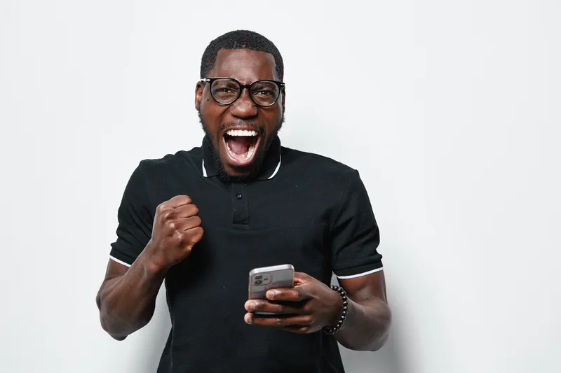 Black man with dark skin and glasses wearing black polo shirt holding smartphone and celebrating success with fist pump and open mouth on white background.