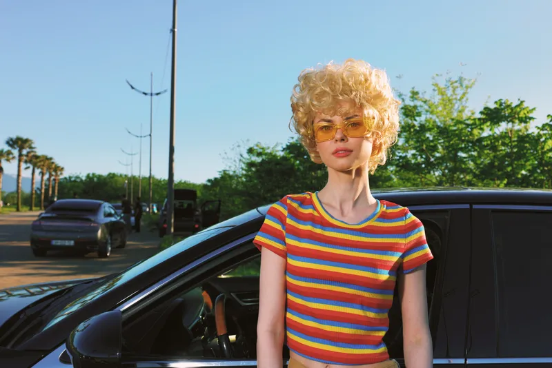 A woman with curly blonde hair in a striped rainbow top leans beside a dark car on a sunny street. Retro film color evokes casual street style and carefree summer vibes, highlighting urban confidence