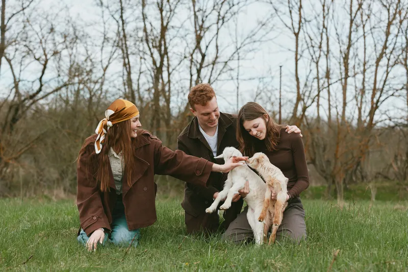 Joyful group outdoors, interacting with a goat kid in a sunny field, casual outfits, natural colors, warm mood, candid lifestyle moment, gentle touch, grassy background, soft breeze, relaxed poses