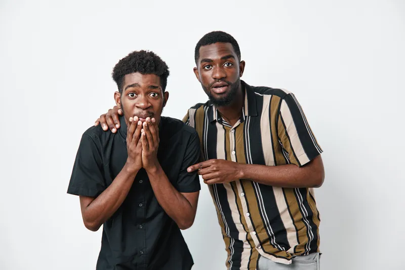 Two young black men with dark skin showing surprise and excitement, one covering mouth and the other pointing, standing against white background in casual clothing.