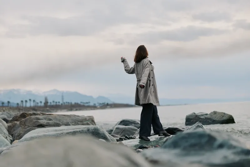 Woman in coat stands on rocks by the sea under cloudy sky. Landscape includes distant mountains and palm trees. Concept of solitude, nature, and calm outdoor moment.