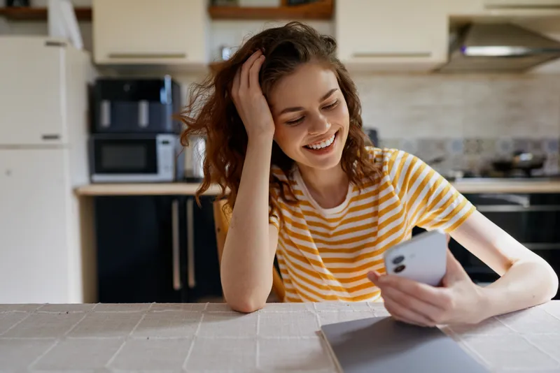 A young woman with brown hair sits at a bright kitchen counter, smiling at her phone while relaxing at home, enjoying a casual moment in a cozy domestic setting.