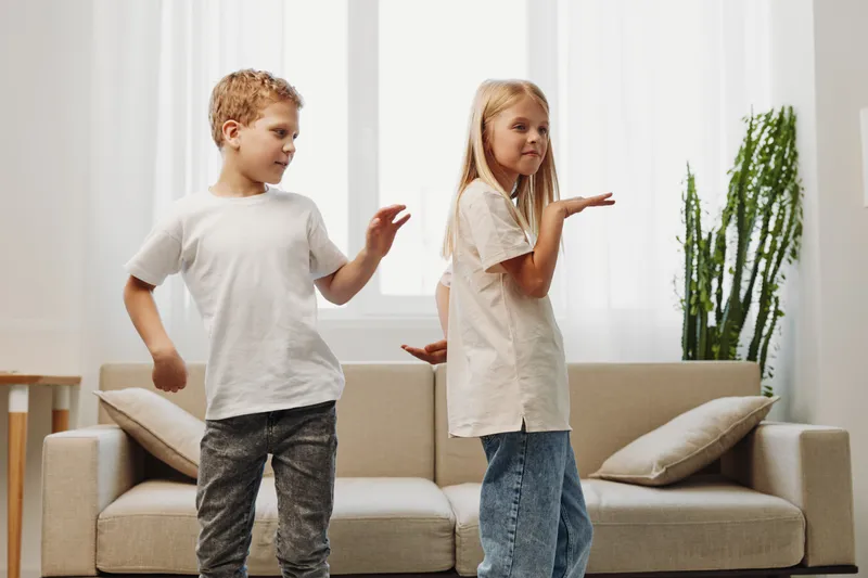 Two children dancing joyfully in casual clothes in a bright living room with modern furniture and indoor plants, expressing happiness and playful energy.