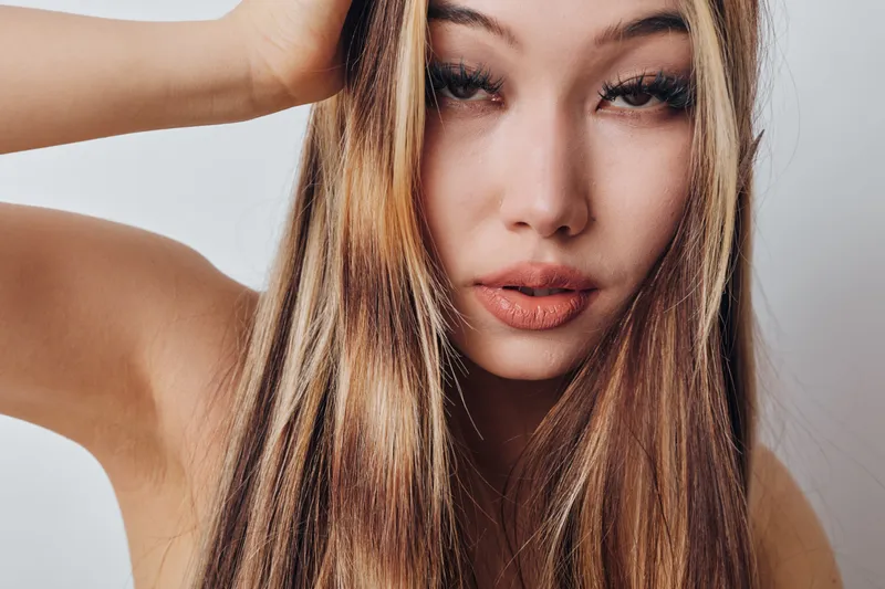 Woman with long hair and natural makeup posing with hand on head against plain background, portrait showing facial features and hairstyle closeup.