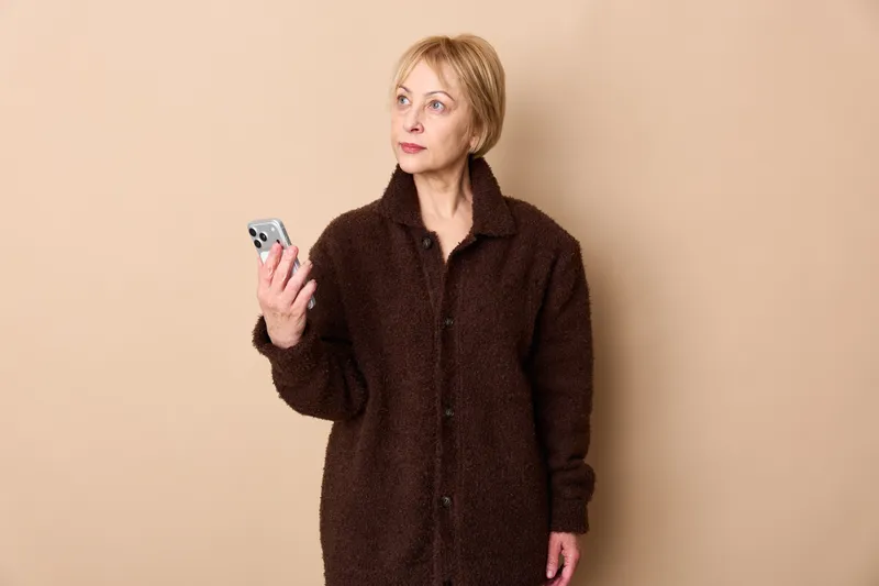 Woman holds dice, showing gambling risk and chance with anxiety, serious expression, beige studio background, uncertainty about decision, senior female portrait