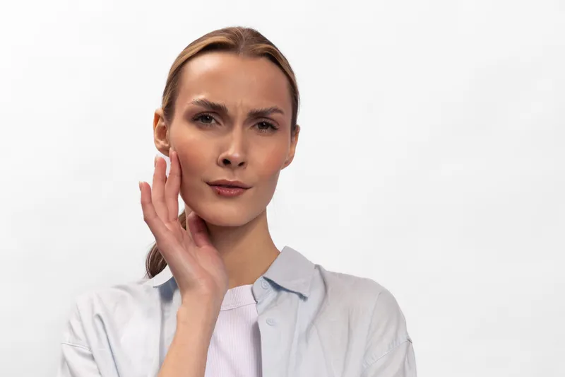 Concerned woman with light skin and blond hair touches her face with a questioning expression. Isolated on white background. Concept for skin care, health, and doubt.