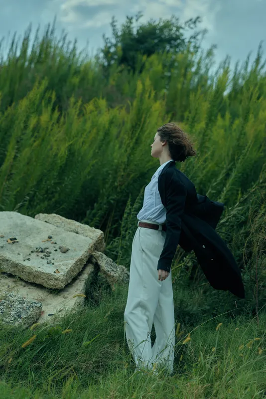 Woman in a flowing coat stands in a grassy field, wind in her hair, tranquil mood, outdoor fashion scene with natural light and dramatic movement.