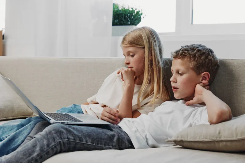 Two focused children, boy and girl, sitting on a sofa watching a laptop screen in a bright living room. Concept of learning, technology use, and childhood curiosity at home.