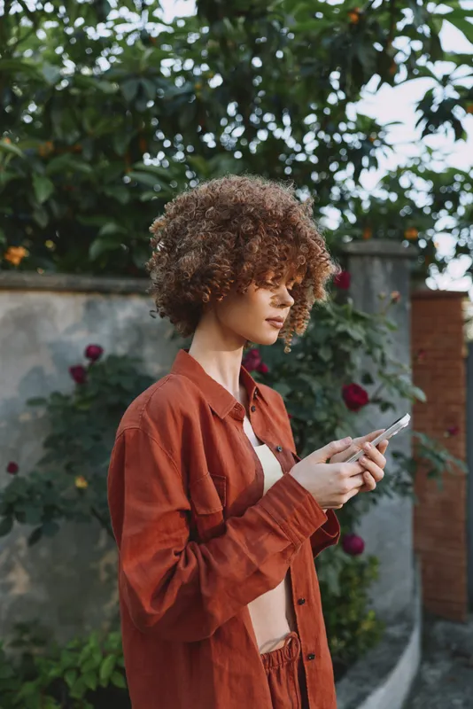 Woman lifestyle outdoors with a smartphone in her hand, wearing a casual jacket and stylish apparel. A bright garden backdrop with greenery and roses creates a warm, confident moment