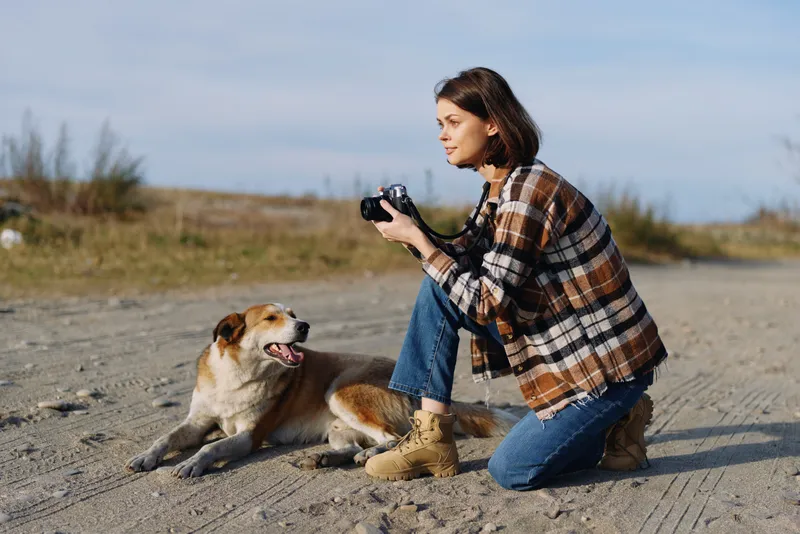 Woman photographer kneels on a sandy beach wearing a plaid jacket and boots capturing a joyful dog with a handheld camera during golden hour outdoor pet portrait and lifestyle moment