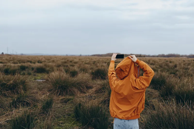 Woman outdoors in a field of grass, orange jacket, cloudy sky behind her, lifestyle portrait, wind lifts the coat, calm solitude, rural nature scene for spring travel