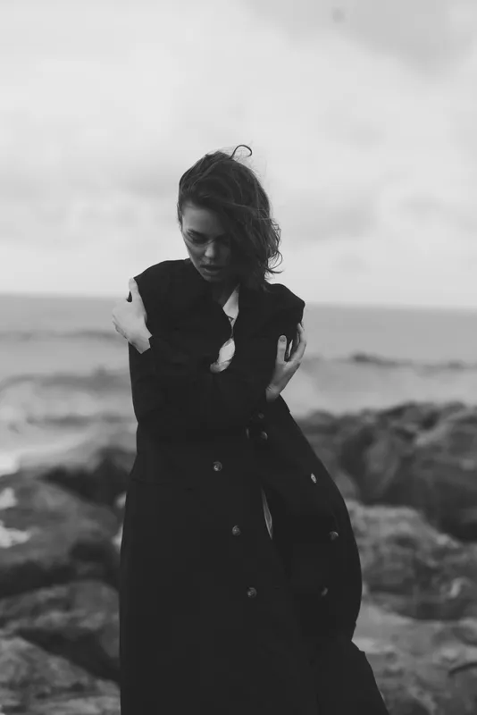 Black and white scene of a woman in a long dark coat embracing herself on a rocky shore, windy atmosphere, moody fashion moment by the sea